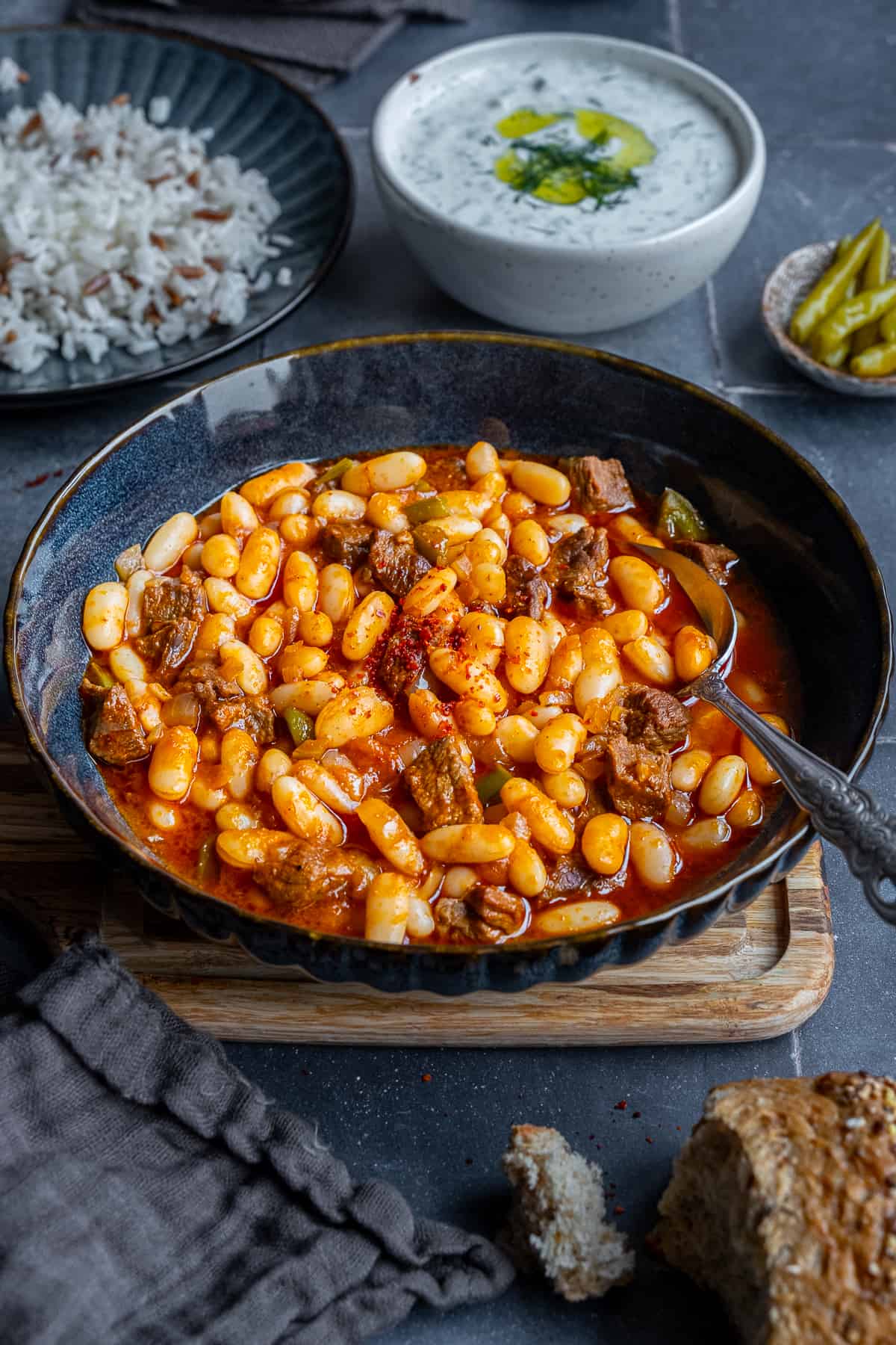 Turkish beans with meat served in a dark colored bowl, Turkish rice, cacik and pickles behind it.