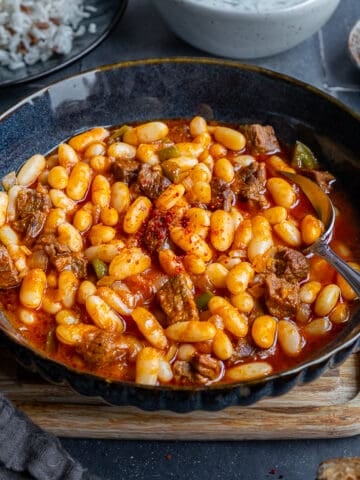 Turkish bean stew in a dark colored bowl and a spoon inside it.