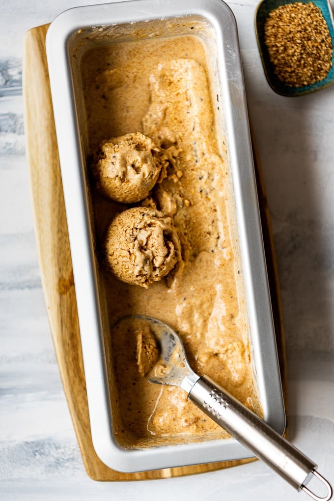 Tahini Almond Milk Ice Cream in a loaf cake tin with an ice cream scoop and sesame seeds on the side.