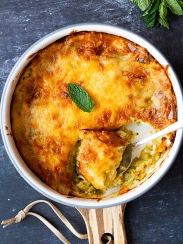 Golden top cheesy zucchini casserole in a white round pan with a spoon in it.