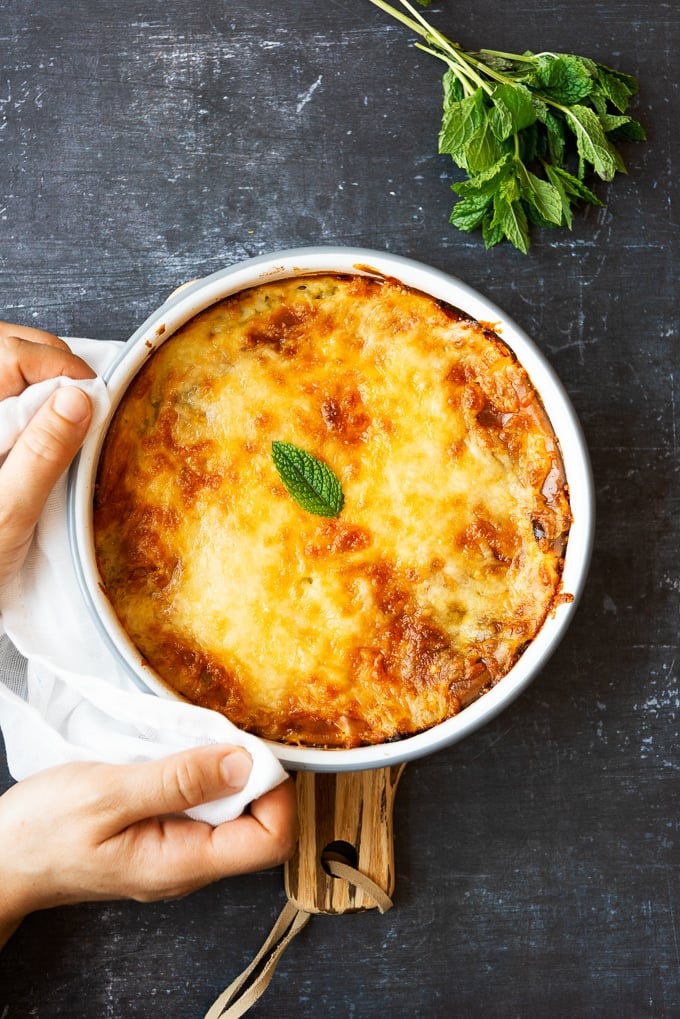 Holding low calorie zucchini casserole with a nicely golden top in a round pan photographed on a dark background.