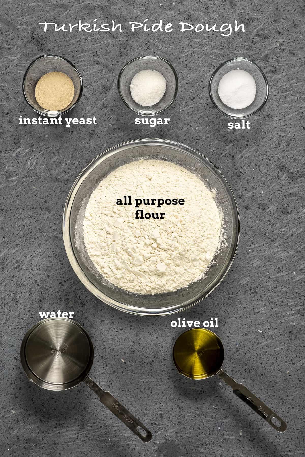 Flour, yeast, sugar, salt, water and olive oil on a grey background.