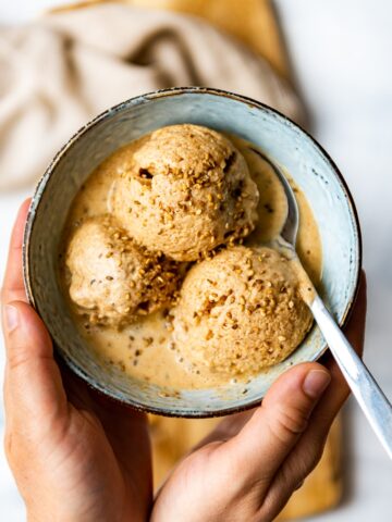 Hands holding a bowl with dollops of tahini almond milk ice cream topped with sesame seeds, a spoon in the bowl accompanies.
