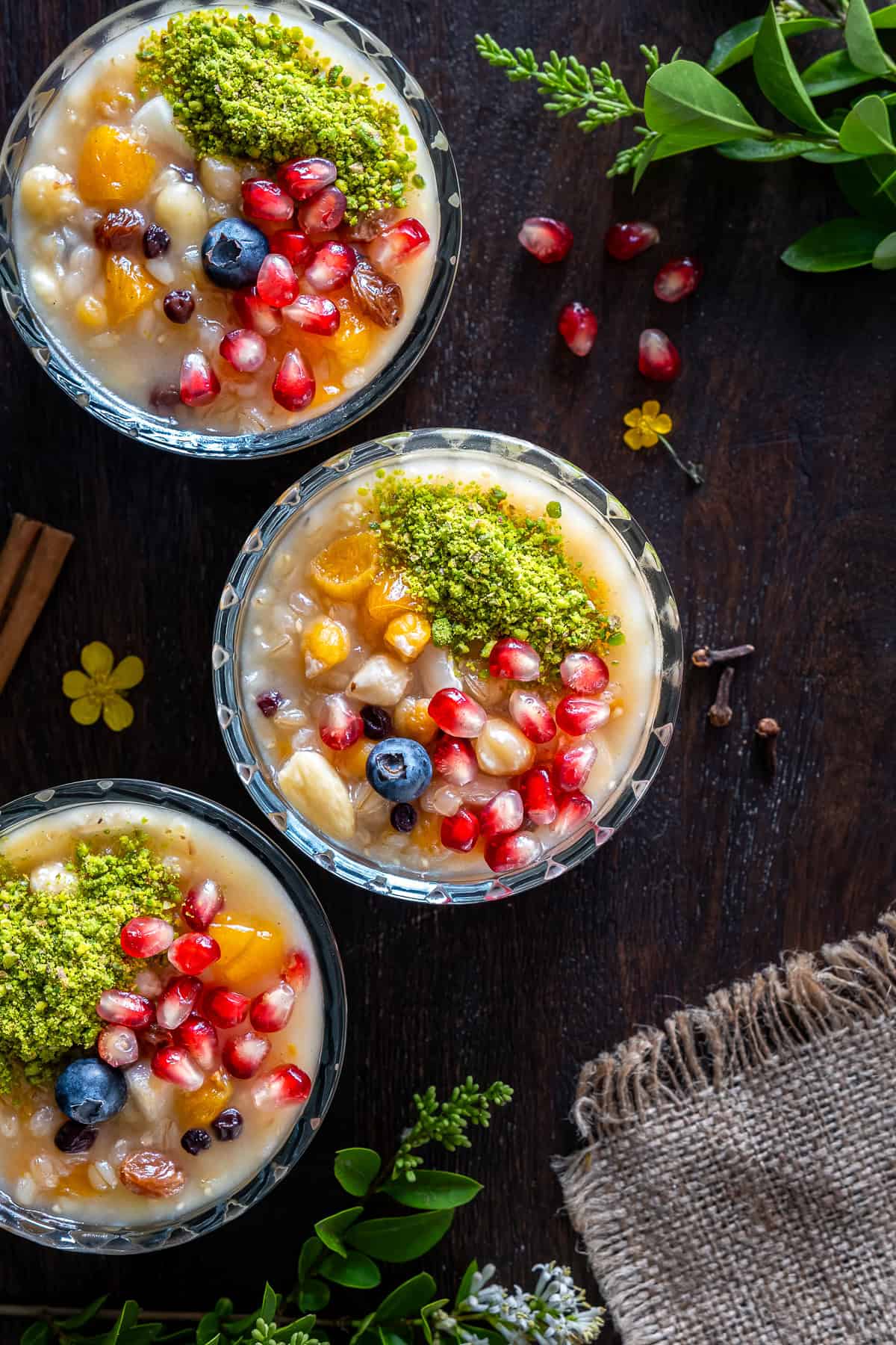 Overhead photo of Turkish ashure (Noah's Pudding) topped with pomegranate seeds, ground pistachios, blueberries, dried apricots, and nuts in glass serving bowls.
