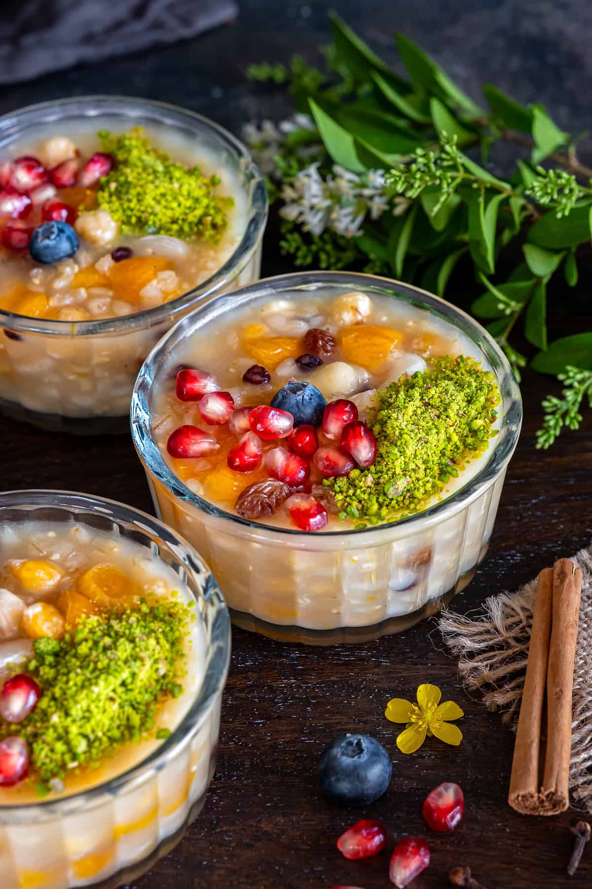 Close-up of Turkish ashure (Noah's Pudding) topped with pomegranate seeds, ground pistachios, blueberries, dried apricots, and nuts in glass serving bowls.
