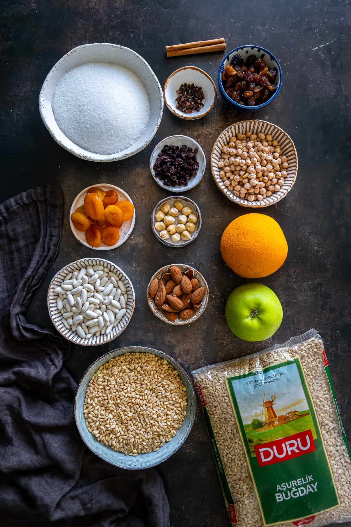 Ingredients for Turkish ashure (Noah's Pudding), including wheat berries, chickpeas, white beans, dried apricots, raisins, currants, almonds, hazelnuts, sugar, orange, apple, cloves, and cinnamon.