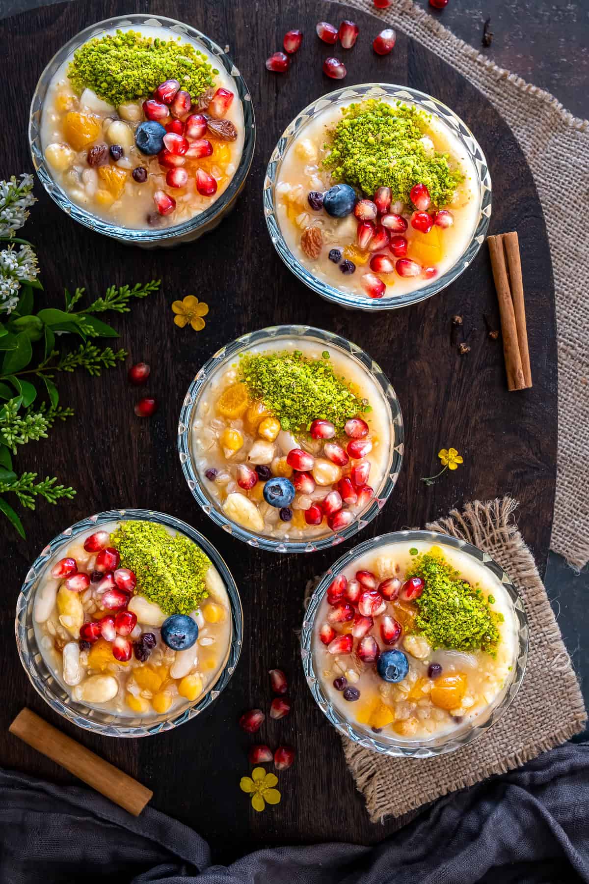 Overhead photo of Turkish ashure (Noah's Pudding) served in individual bowls and topped with pomegranate seeds, ground pistachios, blueberries, dried apricots, and nuts.