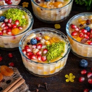 Turkish ashure (Noah's Pudding) topped with pomegranate seeds, ground pistachios, blueberries, dried apricots, and nuts in glass serving bowls.