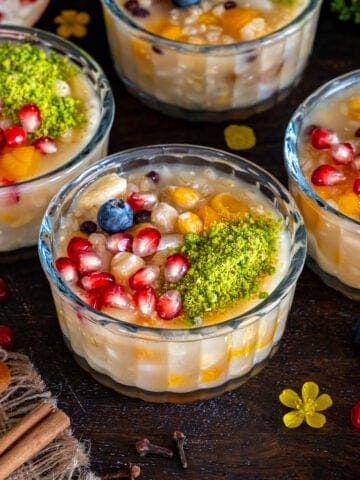 Turkish ashure (Noah's Pudding) topped with pomegranate seeds, ground pistachios, blueberries, dried apricots, and nuts in glass serving bowls.