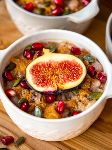 Ashure pudding topped with figs and pomegranate seeds in a white bowl.