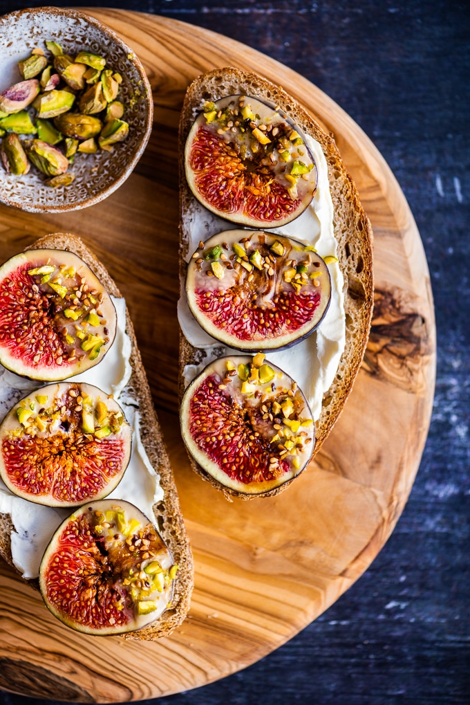 Two bread slices topped with cream cheese and fresh figs dipped coated with tahini and crumbled pistachios on a round wooden board.