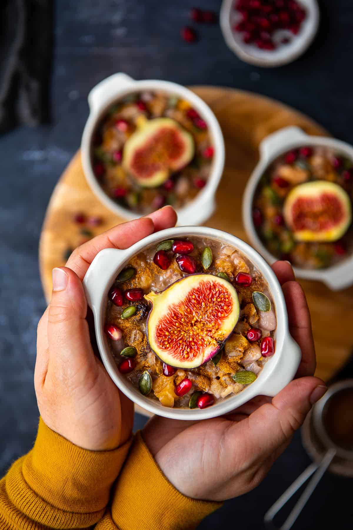 Hands holding a bowl of ashure topped with pomegranate seeds, pumpkin seeds and fresh figs.
