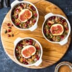 Turkish asure pudding garnished with pomegranate arils, fresh figs and pumpkin seeds in white bowls on a round wooden board.