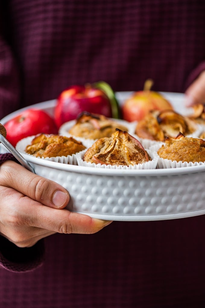 Woman with a claret red sweater holding a white pan full of apple muffins and apples