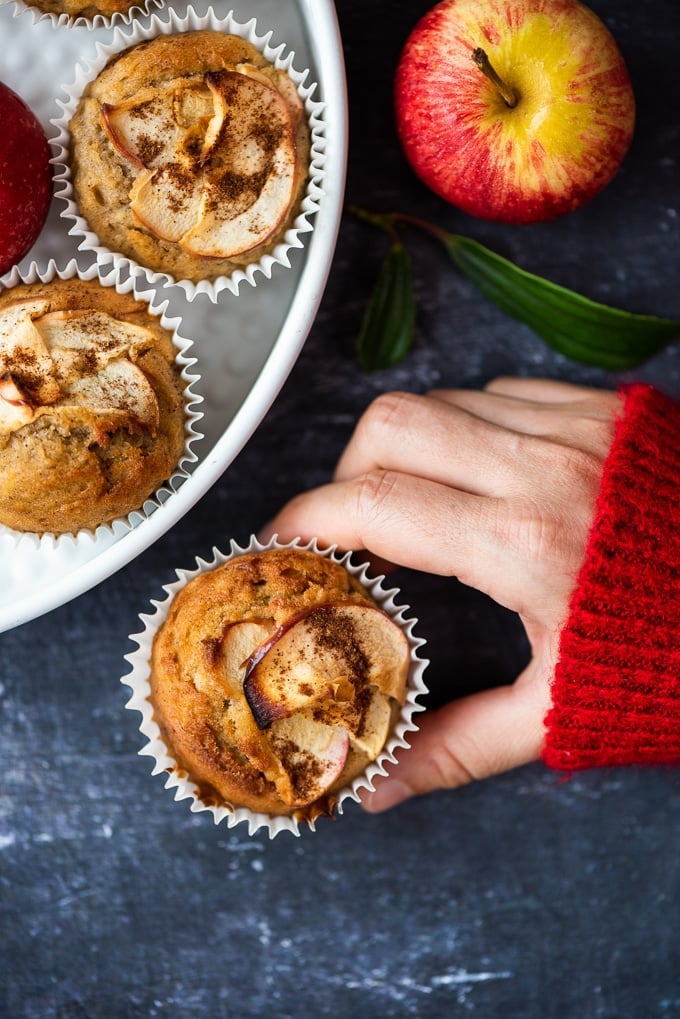 A red sweater wearing woman's hand holding an apple muffin and some other muffins in a white pan, a red apple on the side accompany.