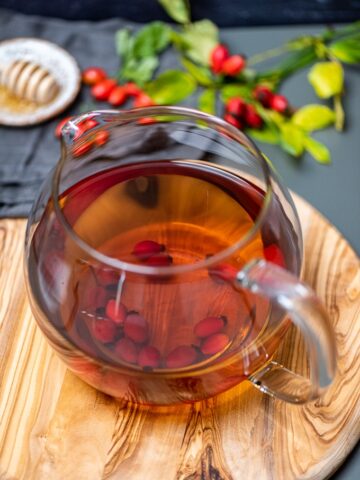 Fresh rose hip tea in a glass teapot on a wooden board, fresh rosehips and honey dipper on the side.