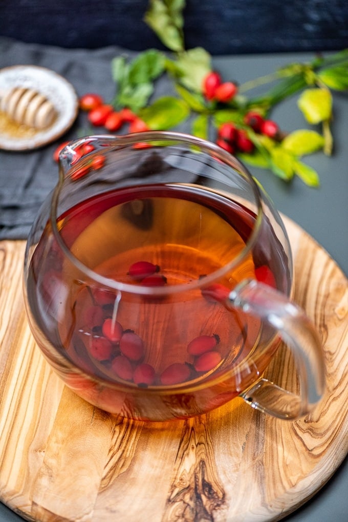 Fresh rose hip tea in a glass teapot on a round wooden board.