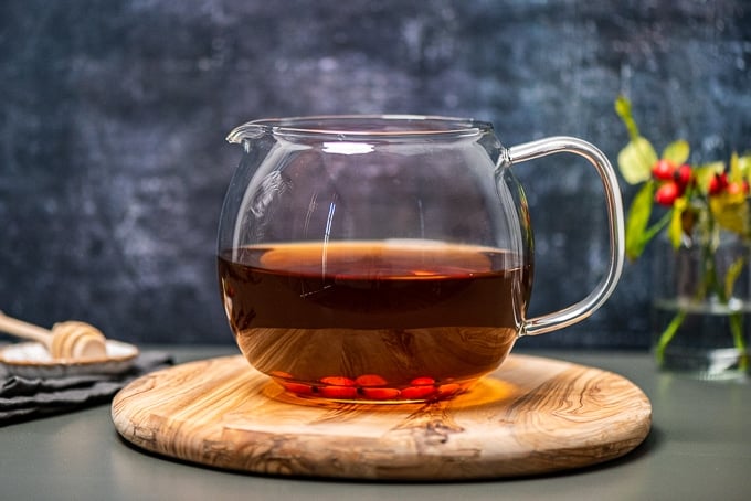 Rosehip tea in a glass pot on a wooden board, honey dipper and fresh rose hips accompany