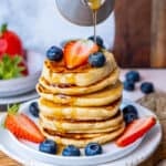 Maple syrup being poured over a stack of pancakes garnished with blueberries and strawberries.