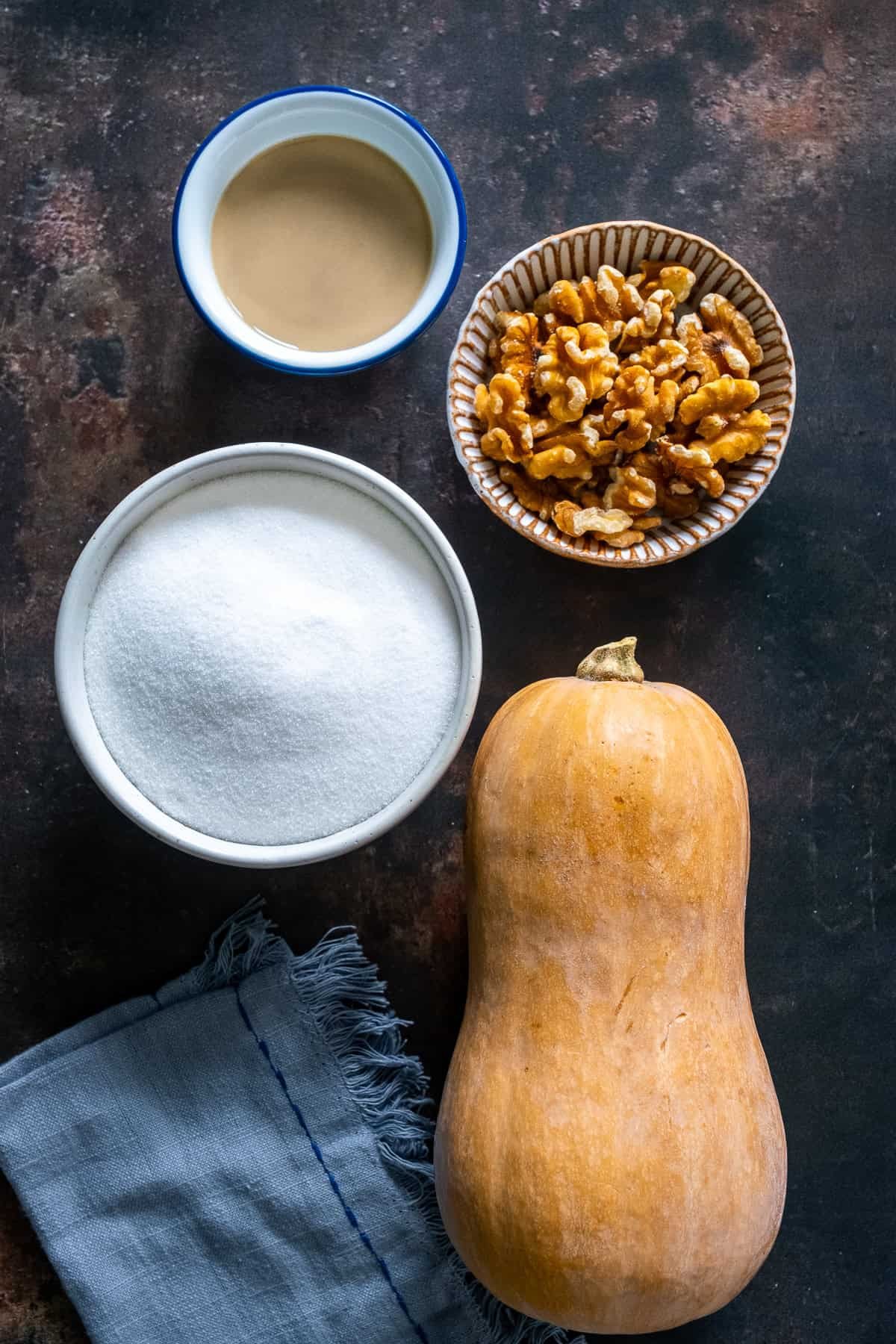 Ingredients for Turkish Pumpkin Dessert (Kabak Tatlısı): a whole butternut squash, a bowl of sugar, a small bowl of tahini, and a bowl of walnuts on a dark background.