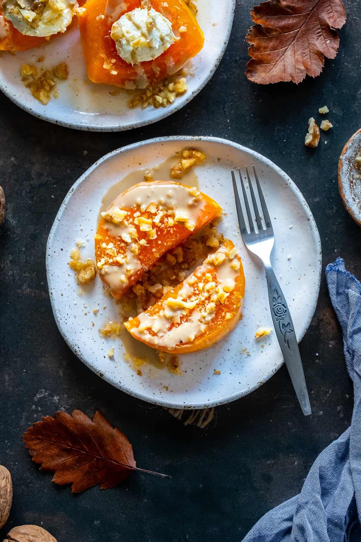 Top view of Turkish Pumpkin Dessert (Kabak Tatlısı) served on white plates, topped with tahini and chopped walnuts, with autumn leaves, walnuts, and a fork on the table.