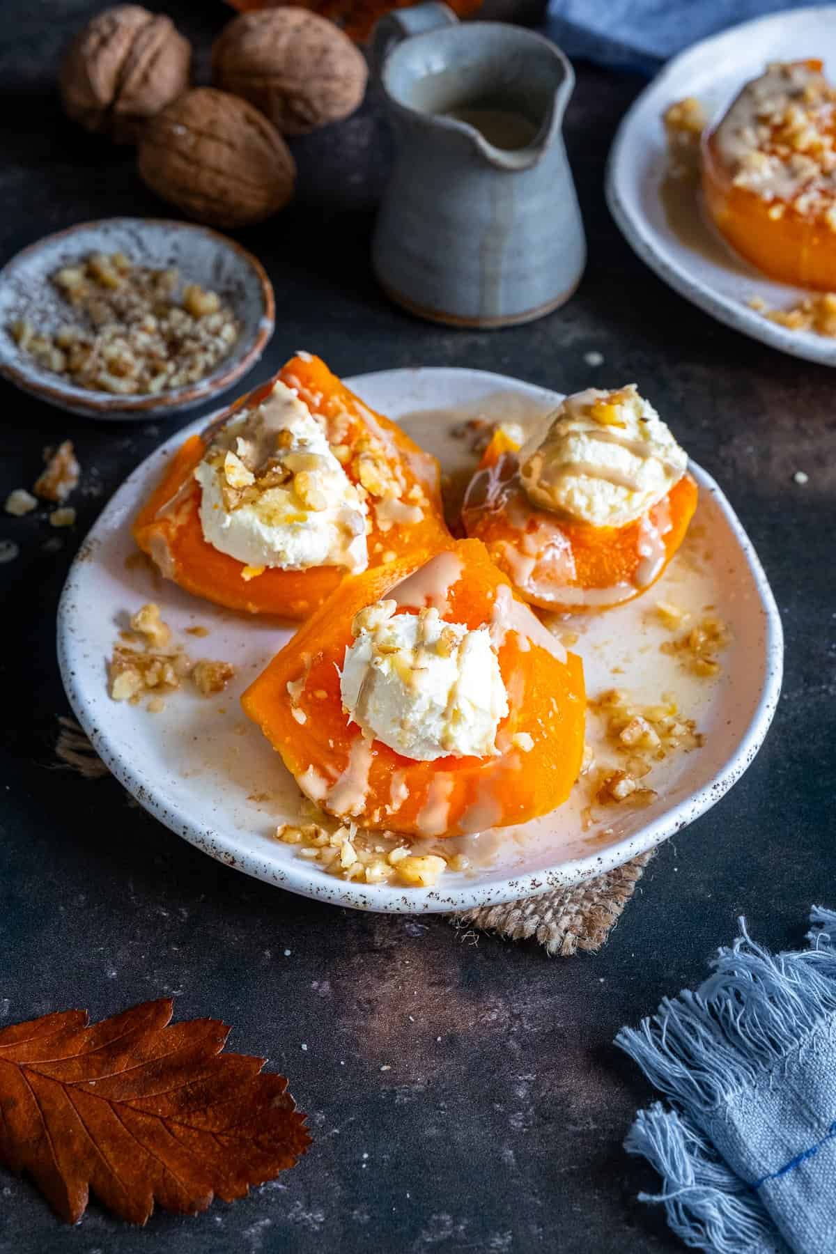 Turkish Pumpkin Dessert (Kabak Tatlısı) on a white plate, topped with clotted cream, tahini drizzle, and chopped walnuts, with walnuts and a jug of tahini sauce in the background.