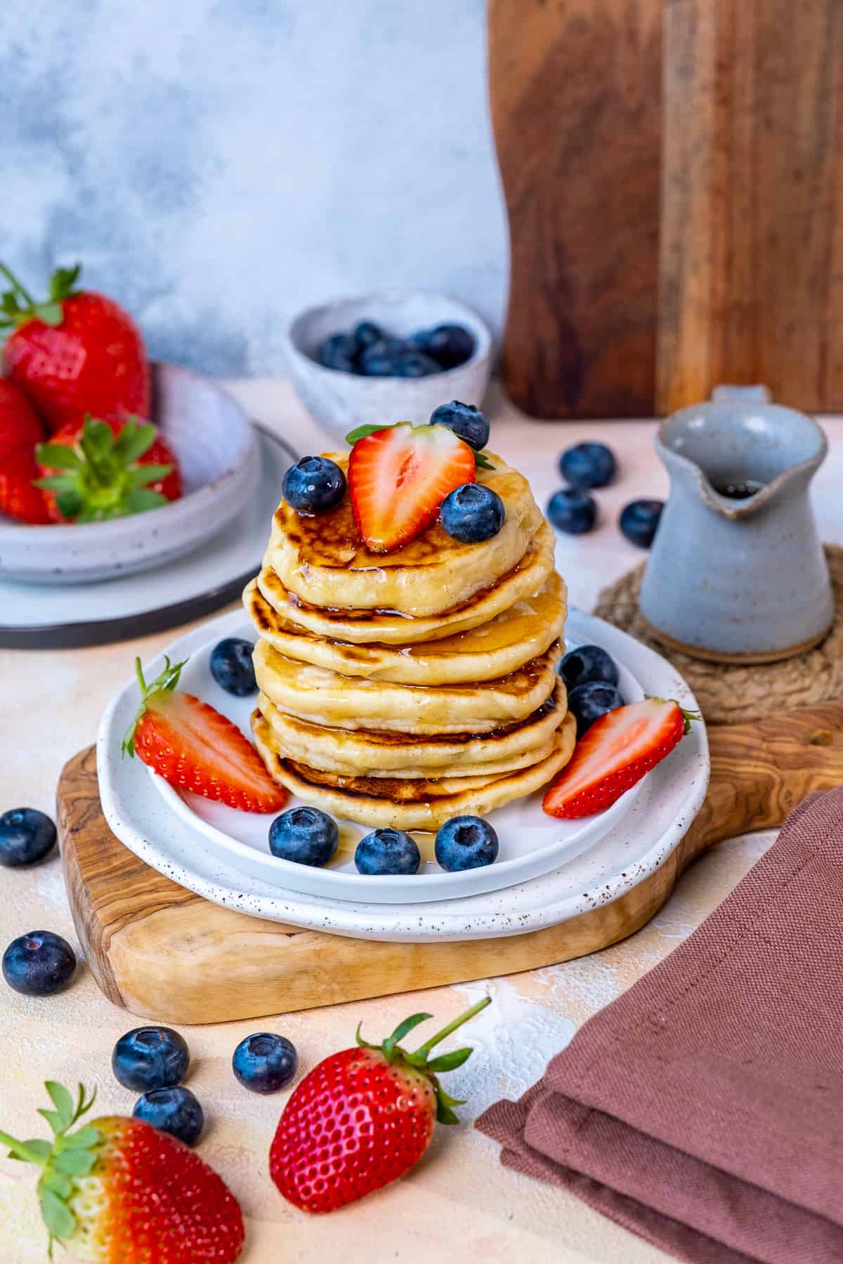 A stack of pancakes served with blueberries and strawberries on a white plate. A small ceramic maple syrup jug and more berries behind them.