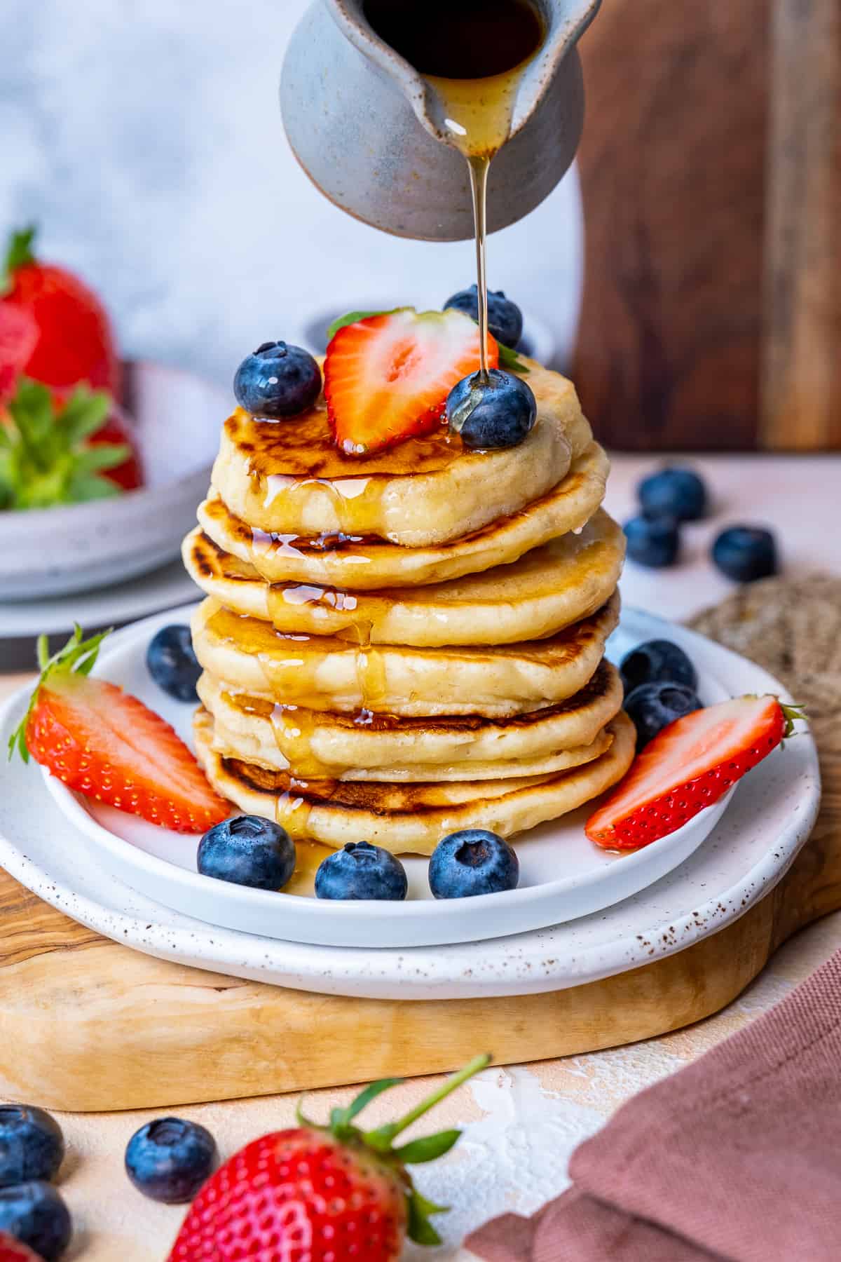 Maple syrup is being poured over a stack of almond milk pancakes served with blueberries and strawberries.