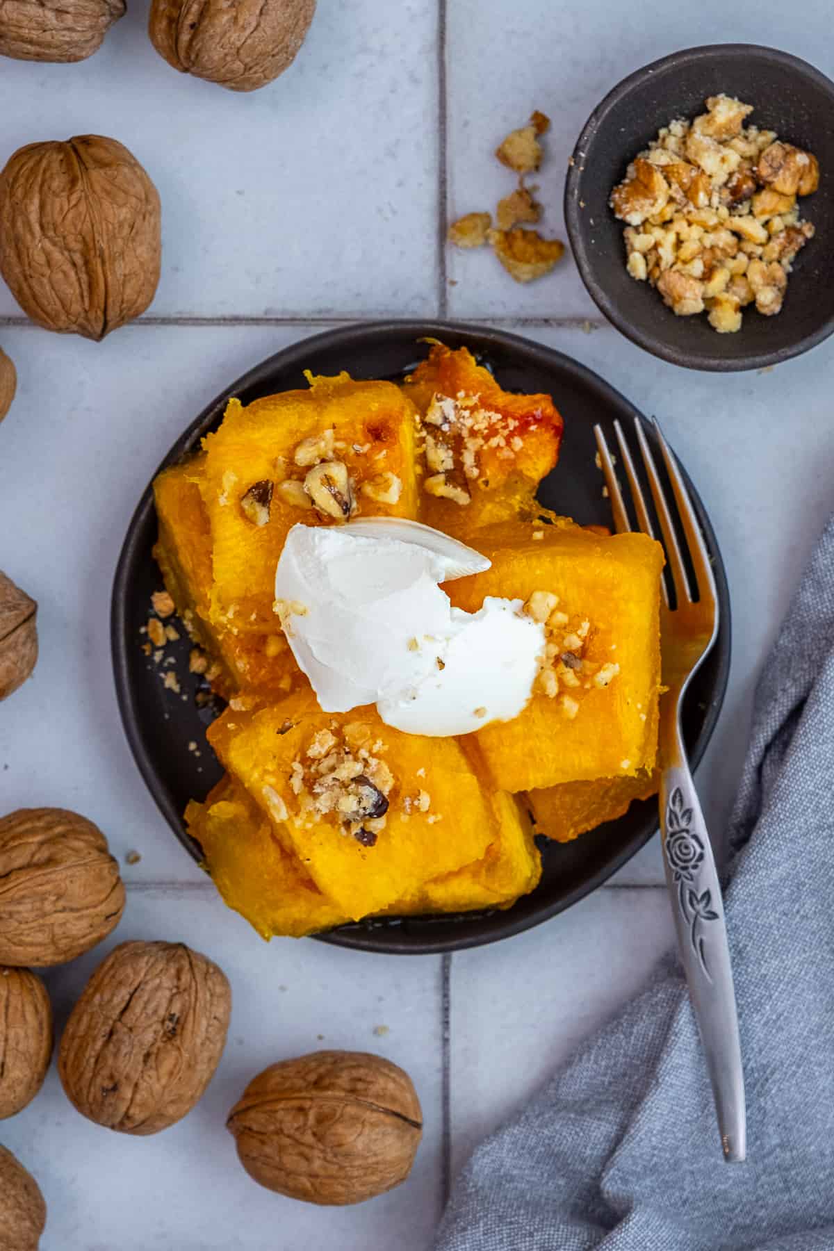 Turkish pumpkin dessert topped with clotted cream and chopped walnuts on a black plate, whole walnuts around it.