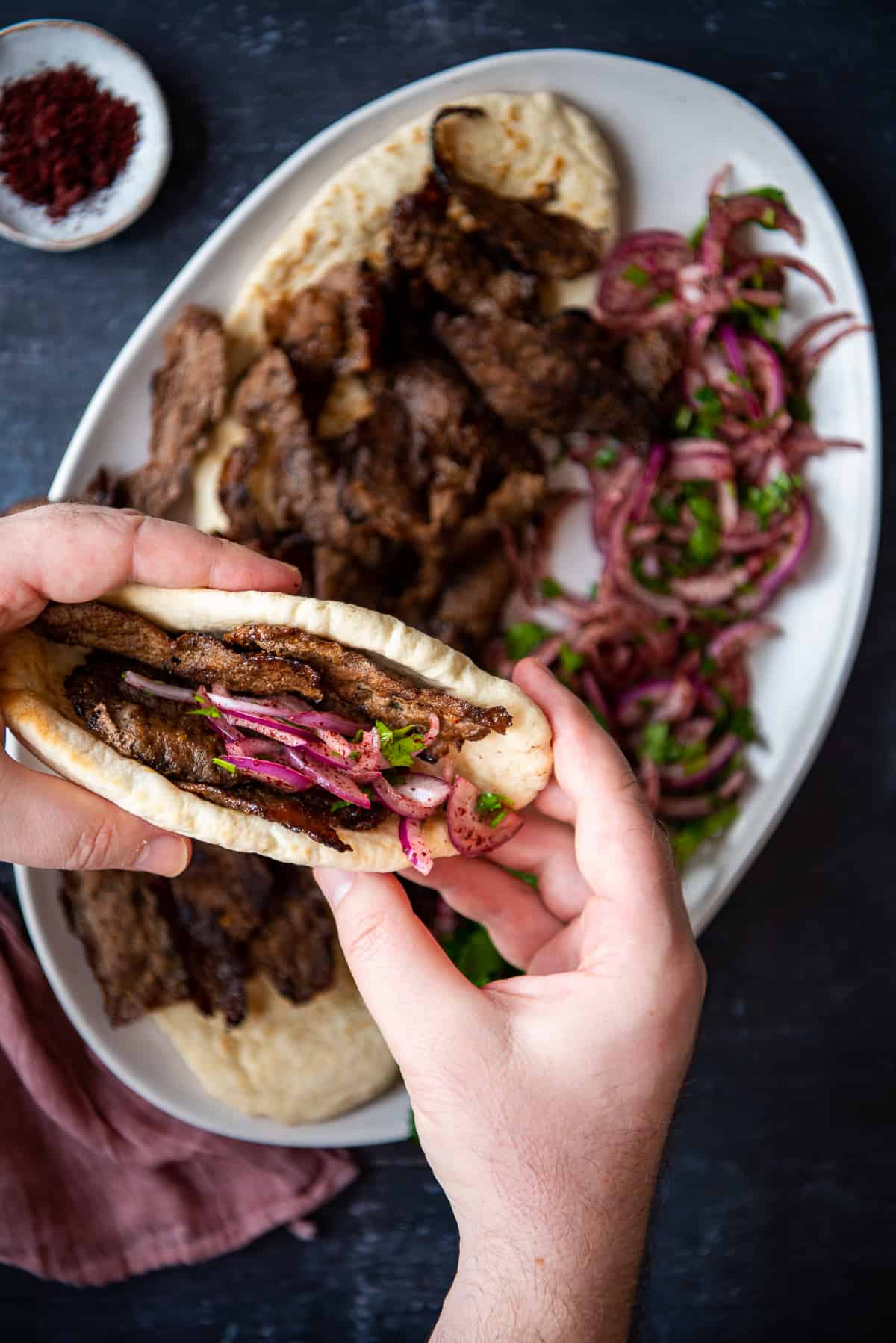 Hands holding a doner sandwich with onions over a plate loaded with more doner slices.