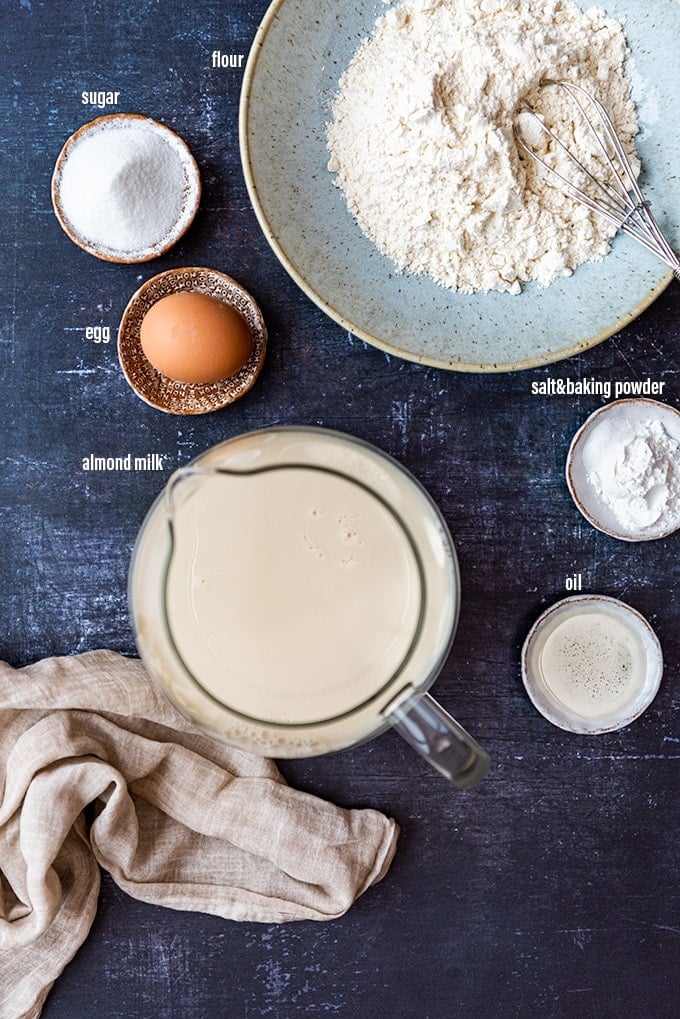 Almond milk in a jug, egg, oil, flour, sugar and baking powder in bowls photographed on a dark background.