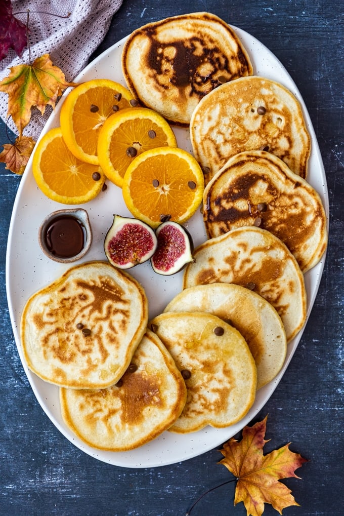Almond milk pancakes served with orange slices, figs and maple syrup on a white oval plate.