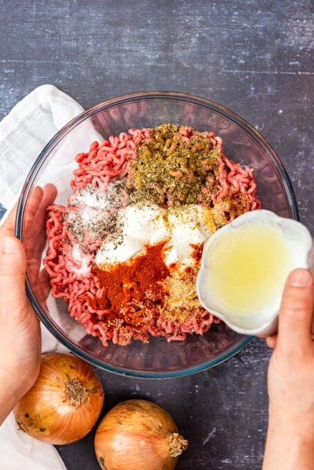 A hand pouring onion juice over ground beef doner meat ingredients in a large glass bowl.