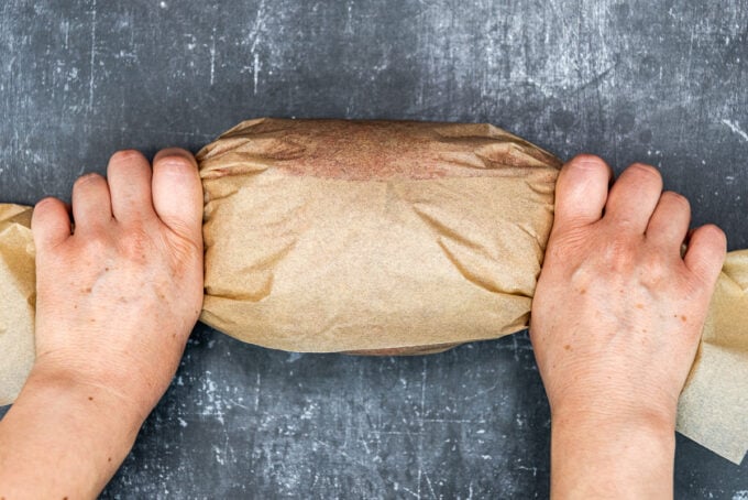 Hands tightening a log of beef doner that is wrapped with brown baking paper from two sides.