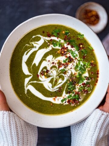 Woman with white jumper sleeves holding vegan spinach soup with dairy free yogurt topping in a big white bowl.