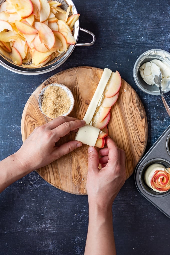 Hands rolling a puff pastry strip up filled with apple slices and cream cheese inside on a circular wooden board.