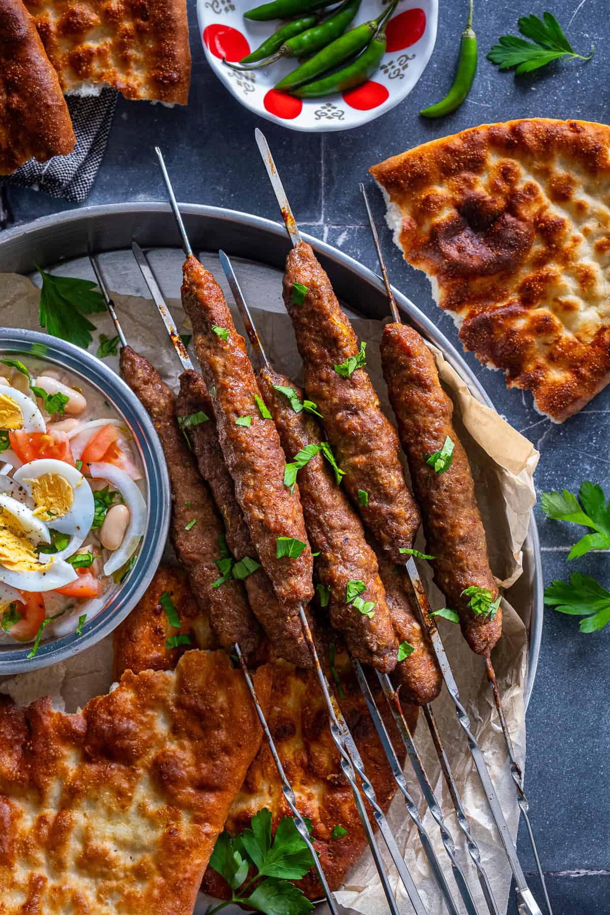 Overhead view of Turkish şiş köfte (meatball skewers) on a metal platter with pide bread, white bean salad topped with boiled eggs, fresh parsley and green peppers.