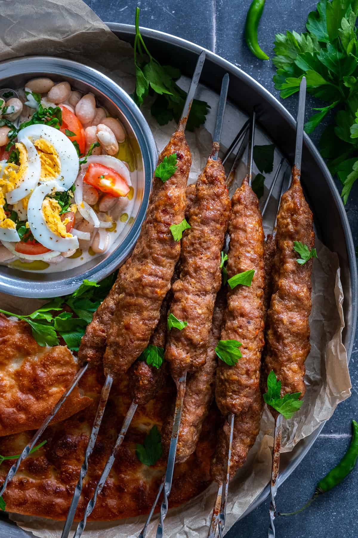 Turkish şiş köfte (meatball skewers) garnished with fresh parsley and served with pide bread and Antalya-style white bean salad topped with boiled eggs.