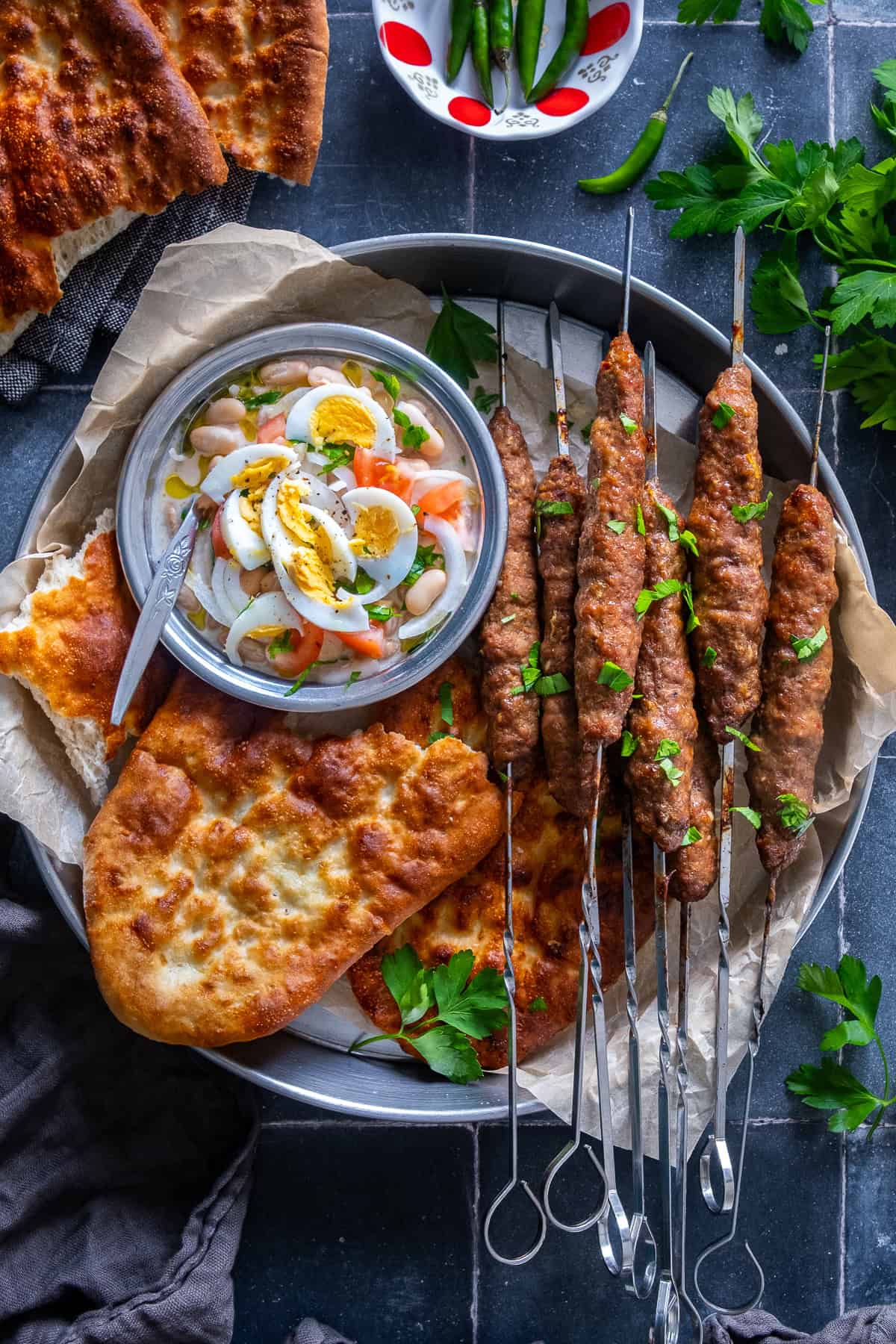Overhead view of Turkish şiş köfte (meatball skewers) served on a metal platter with pide bread, Antalya-style white bean salad topped with boiled eggs, fresh parsley and green chili peppers.