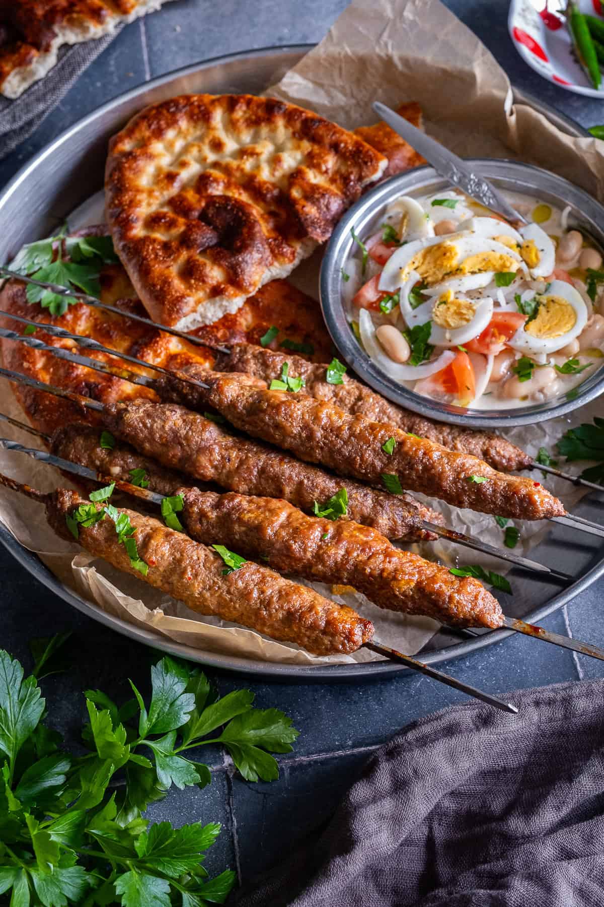 Turkish şiş köfte (meatball skewers) garnished with parsley and served on a metal platter with golden pide bread and Antalya-style white bean salad topped with boiled eggs.