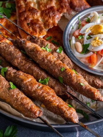 Turkish şiş köfte (meatball skewers) served on a platter with pide bread, white bean salad topped with boiled eggs, and fresh parsley.