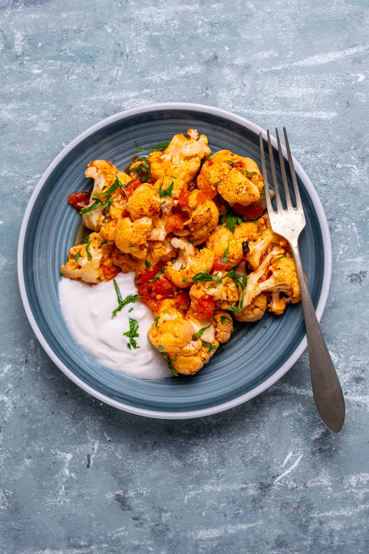 Baked cauliflower dish served on a bluish plate, yogurt sauce and a fork on the side.