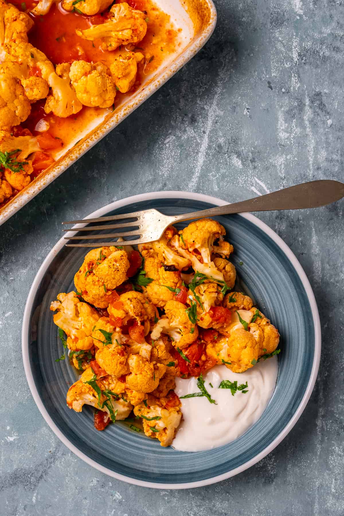 Baked cauliflower with a tomato sauce served on a blue plate, a fork and some yogurt on the side.