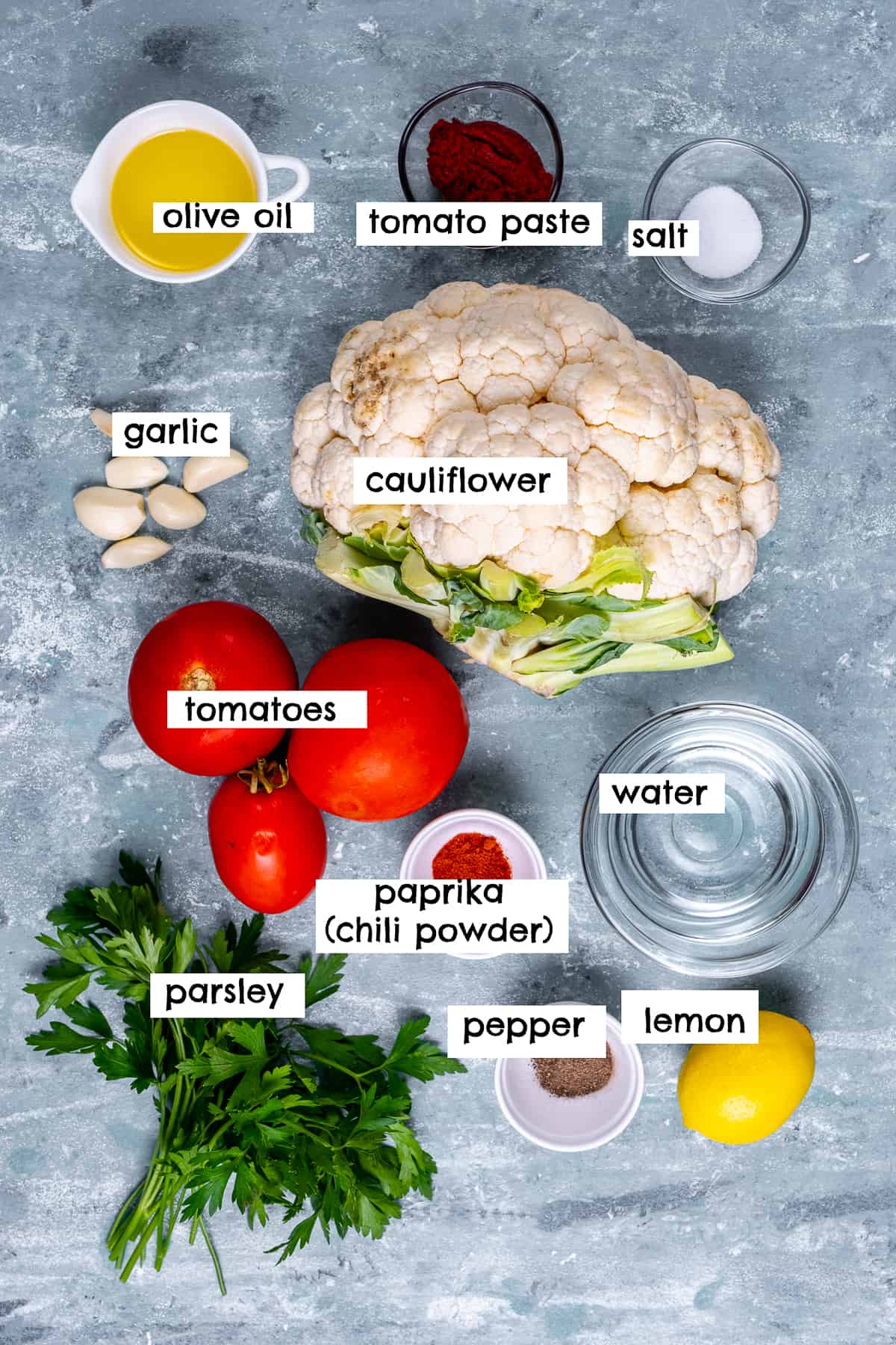 A head of cauliflower, tomatoes, lemon, olive oil, garlic, tomato paste, parsley, salt, pepper and paprika photographed on a light grey backdrop.