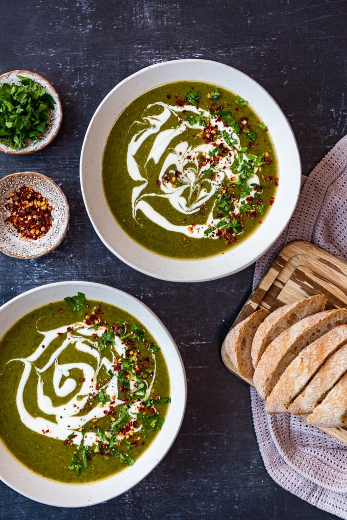 Two bowls of spinach soup with lentils, red pepper flakes, chopped parsley and bread slices on a dark background