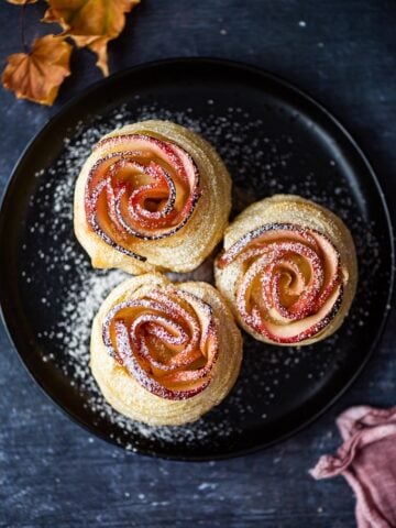 Puff pastry apple roses dusted with powdered sugar on a black plate, fall leaves on the side.