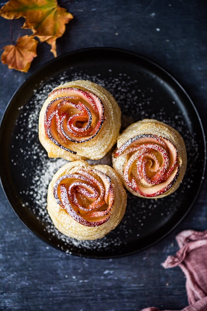 Cream cheese puff pastry apple roses with powdered sugar on a black plate.