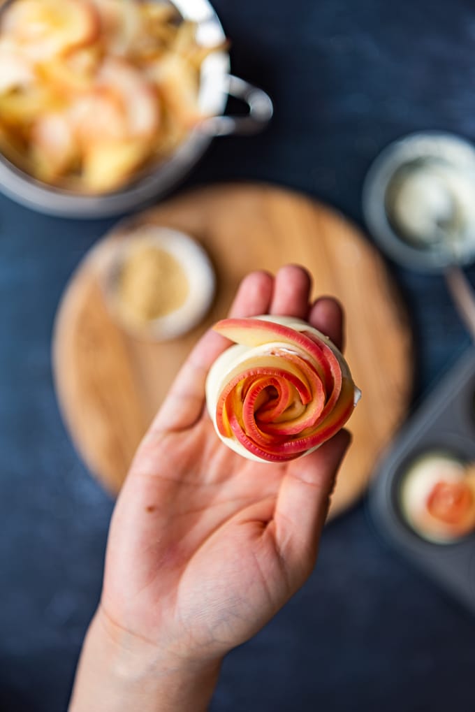 A hand holding a puff pastry strip with apples rolled into a rose.