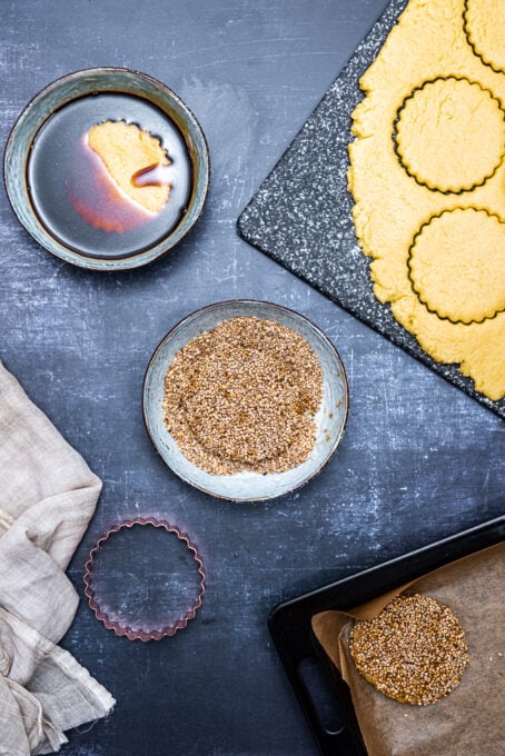 Cookies cut in round shape, dipped into a bowl of molasses and another cookie dough in another bowl of sesame seeds.