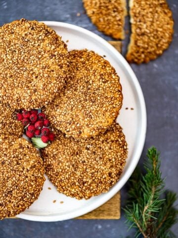 Turkish tahini sesame cookies on a white plate, holly berries in the middle and pine branch on the side.